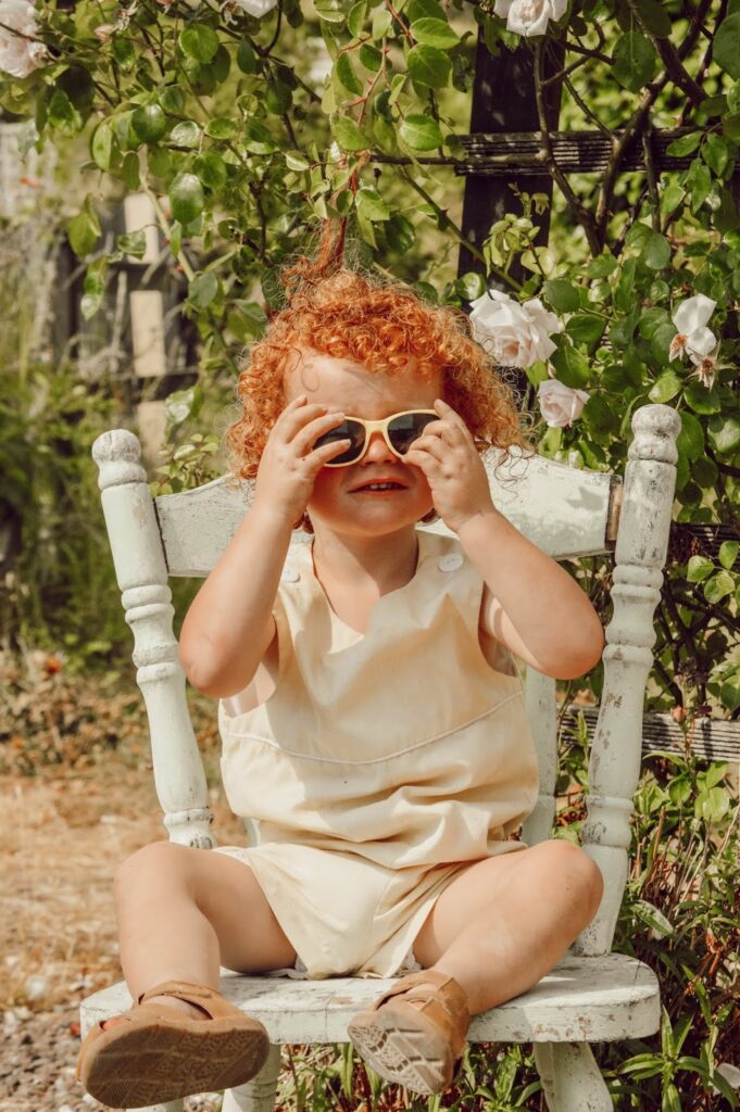 Red haired child sitting on a chair wearing sunglasses and a yellow romper in sunny weather