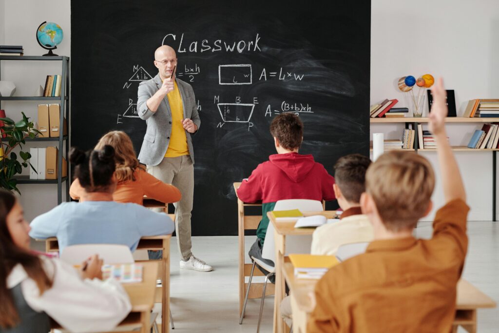 School classroom with a student raising his hand in the air and a teacher pointing at him in front of a blackboard