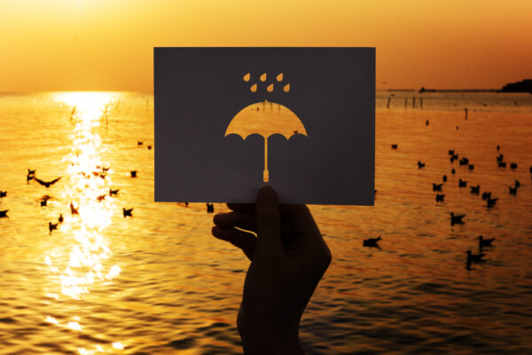 Silhouette of a hand holding up a piece of black cardboard with an umbrella cut out over the sea at sunset