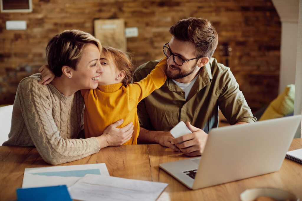 Daughter kissing her mother while sitting with father and a laptop at a table