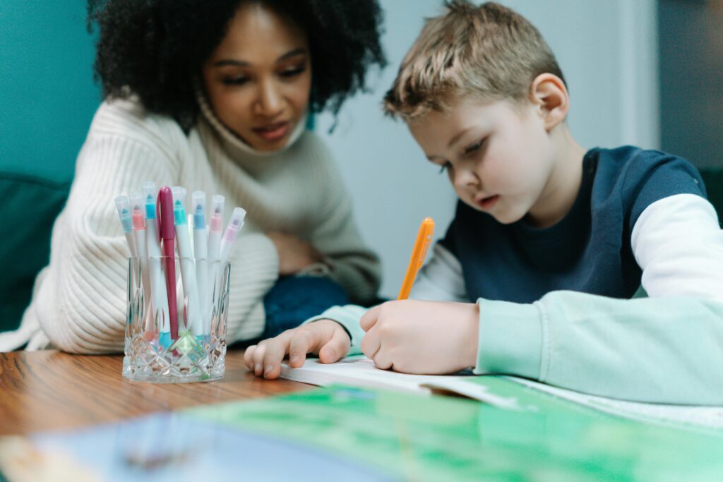 Woman with brown curly hair teaching a boy with blonde hair how to write