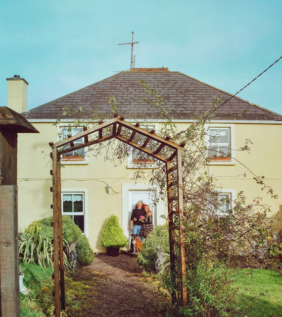 Man, woman and dog standing outside a yellow farmhouse