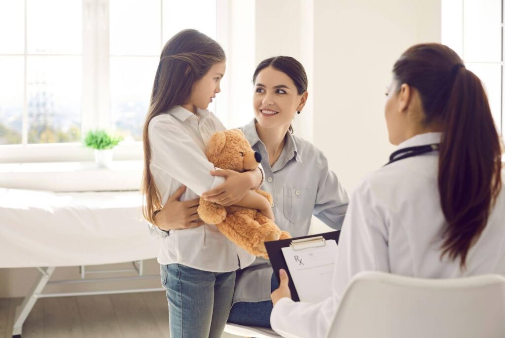 Young girl holding a teddy bear speaking with doctor and mother
